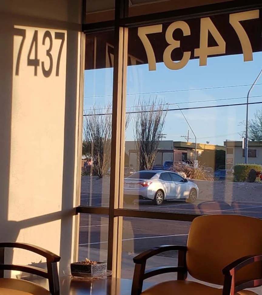 Looking out through the front window of the Arizona Computer Guru office at golden hour: the street number 7437 on the wall inside and again reversed on the glass, waiting-room chairs in the foreground, and ocotillo and low desert buildings across the parking lot.