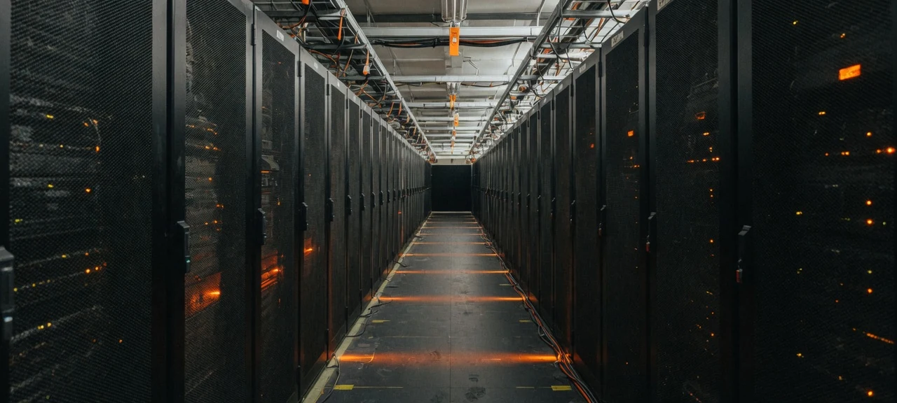 A datacenter cold aisle of tall perforated server cabinets receding to a vanishing point, restrained orange status LEDs and floor lighting in the dark.