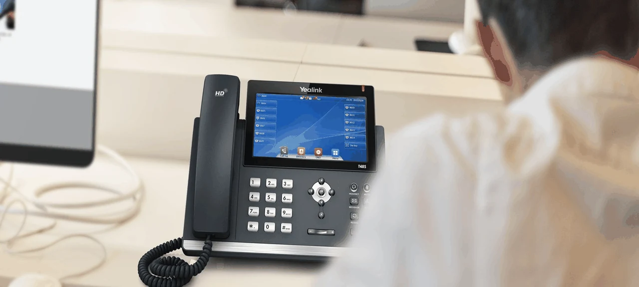 A Yealink T48S IP desk phone on a desk in a busy open-plan office, its keypad and function keys clearly visible and its color display lit with line keys, a colleague working at the next desk behind it.